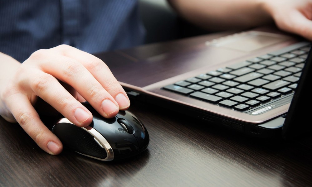 A person's hand using a mouse with a laptop, with their other hand in the background.