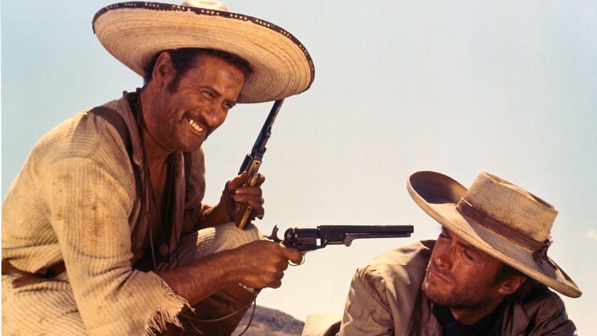 Eli Wallach como Tuco sonriendo y sosteniendo dos pistolas, mientras que Clint Eastwood como Blondie lo mira con fastidio en El bueno, el malo y el feo.