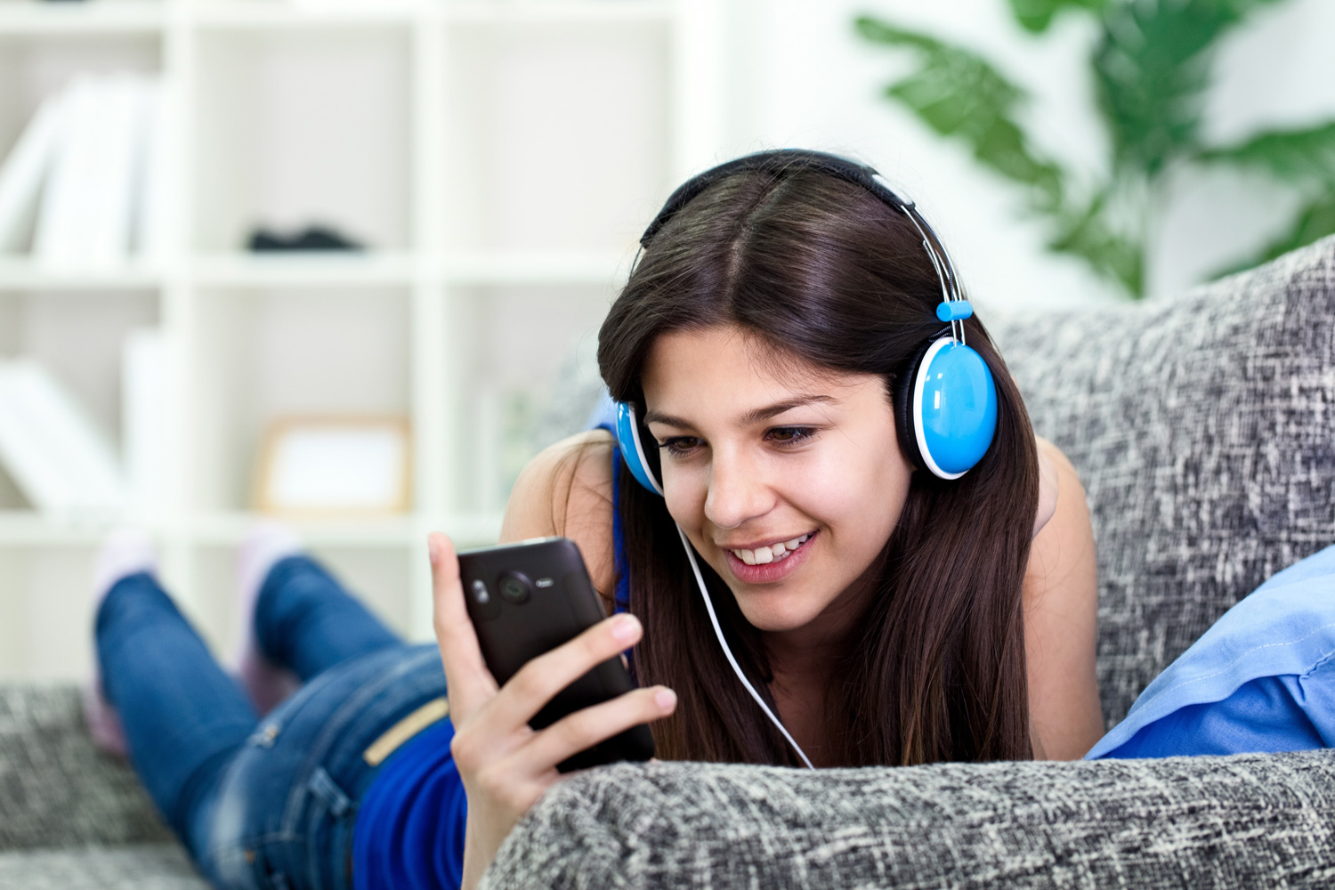 Eine Frau mit Kopfhörern schaut auf ein Smartphone, während sie auf einem Sofa Musik hört.