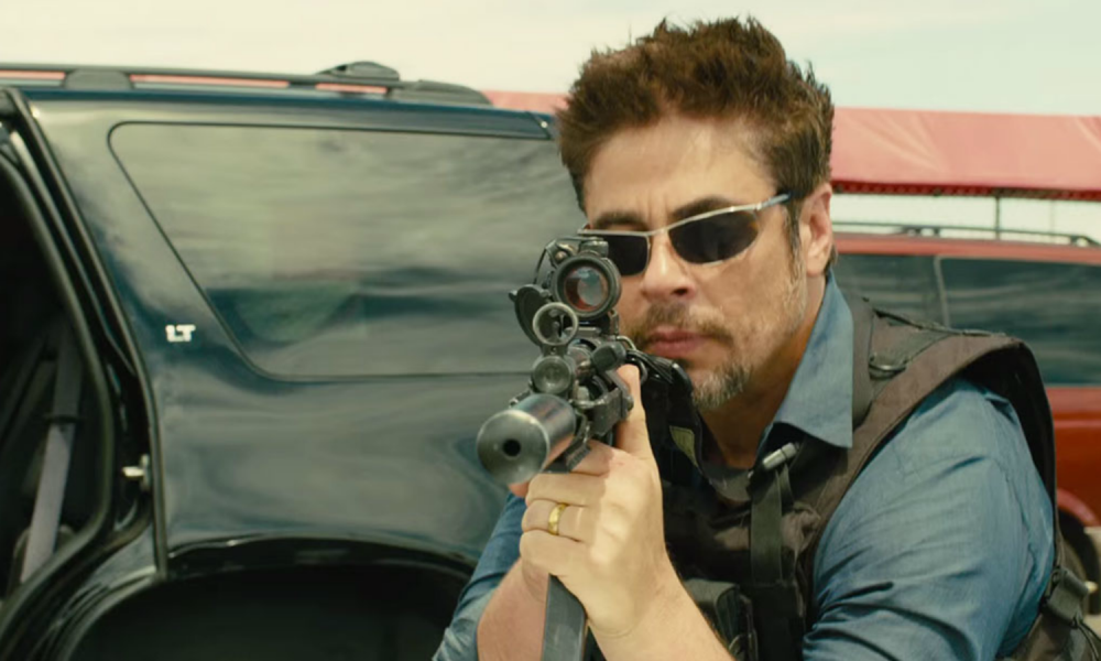 Bencio del Toro points a gun and stares.