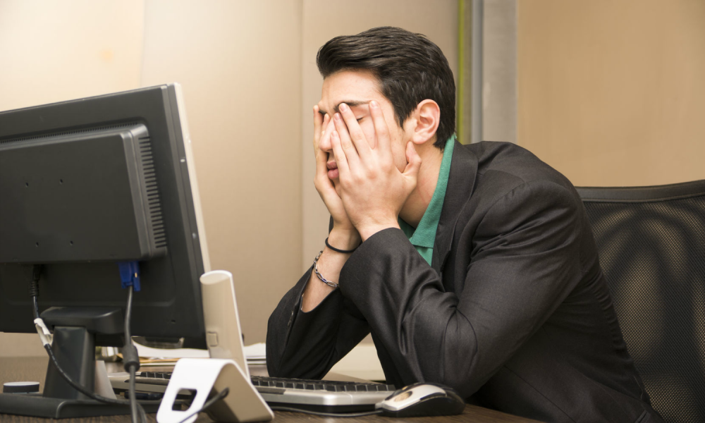 A person sitting at a desk, in front of a computer monitor with their head in their hands.