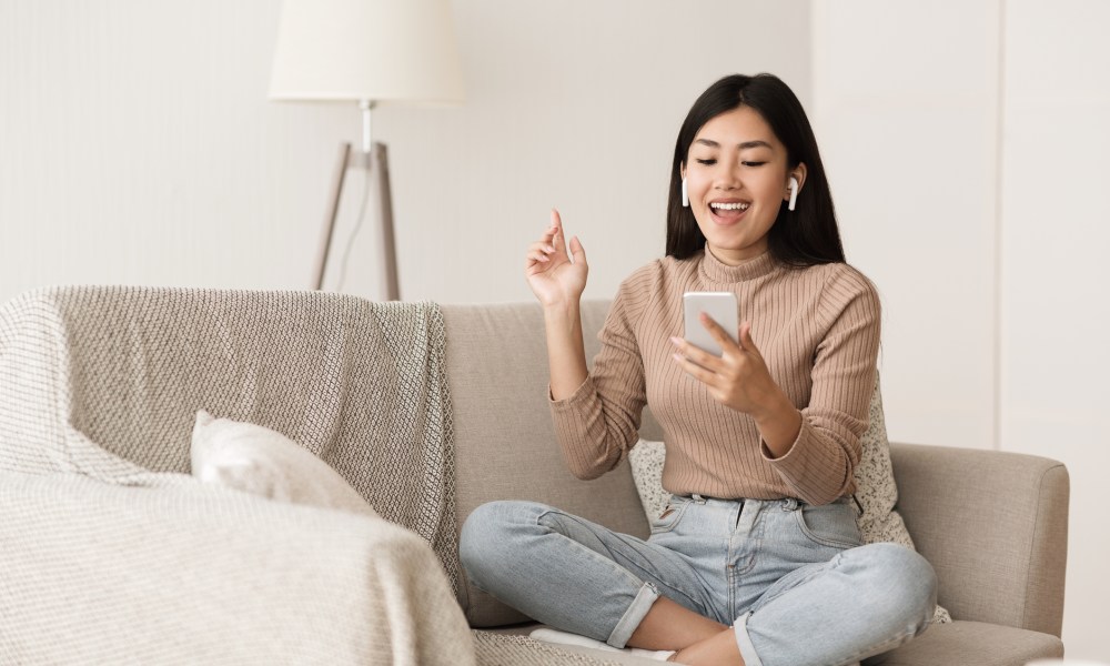 A woman sitting on a couch, wearing airpods and holding and looking at a smartphone.