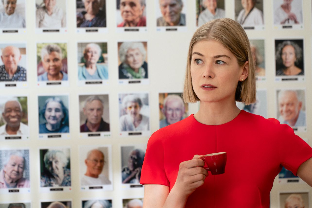 Rosamund Pike hält eine Tasse Kaffee vor einer Wand voller Fotos.