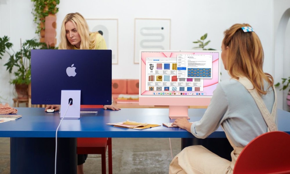 Two people use iMacs on a desk in an office.