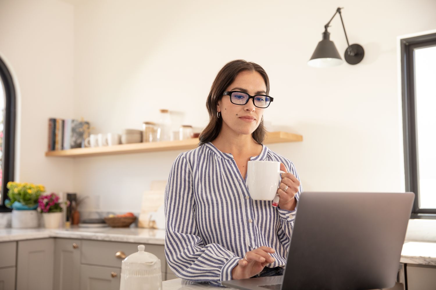 Uma mulher usando armações de eco, sentada em frente ao computador, tomando café.