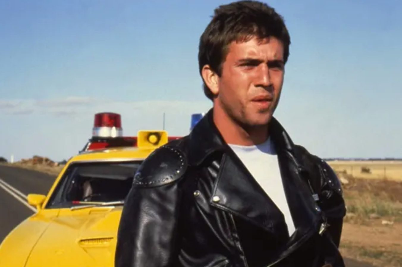 A man in a leather jacket sits on the front of a car.
