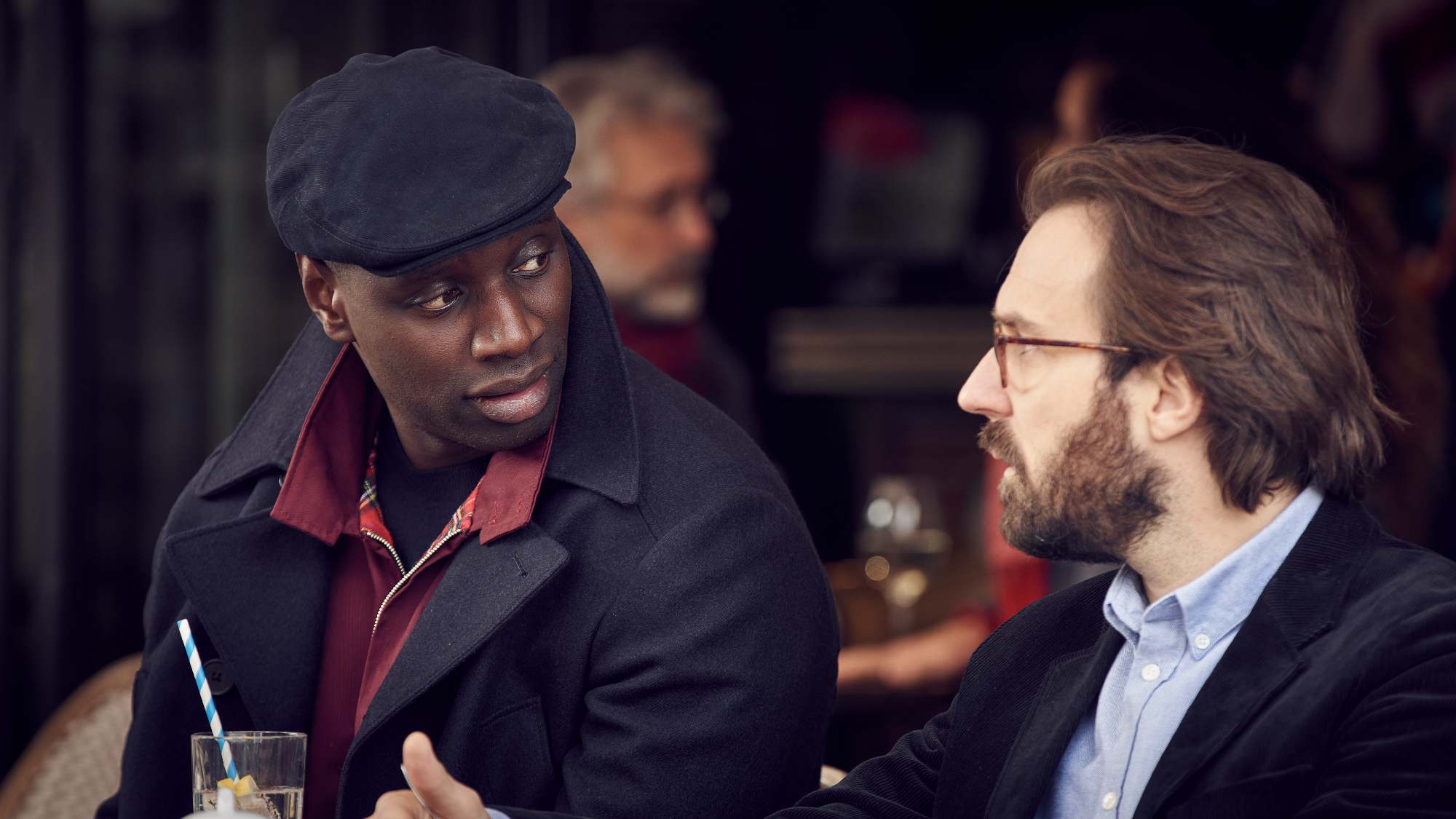 Omar Sy in Lupin sitting on a patio with someone having a conversation.