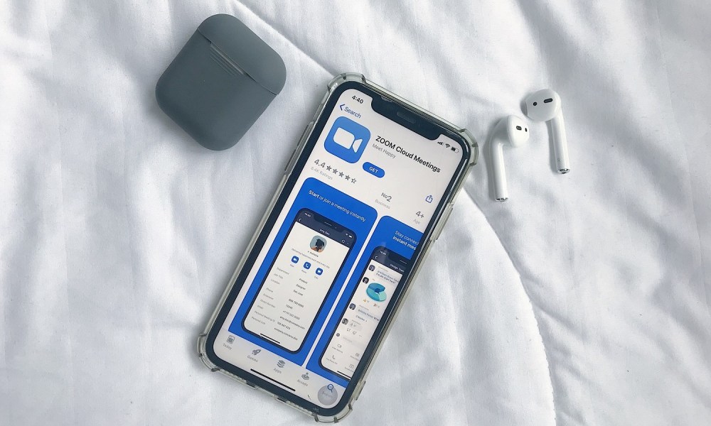 An iPhone with the Zoom mobile app's app store listing on its screen. AirPods and their case are nearby, all on a white fabric background.