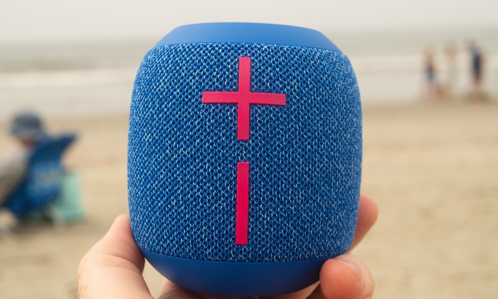 A hand is holding the Wonderboom 3 speaker in front of a beach background.