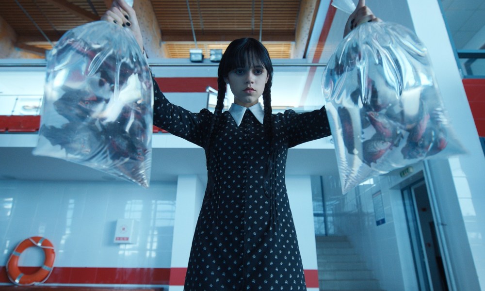 Jenna Ortega, as Wednesday Addams, holds two bags filled with piranhas.