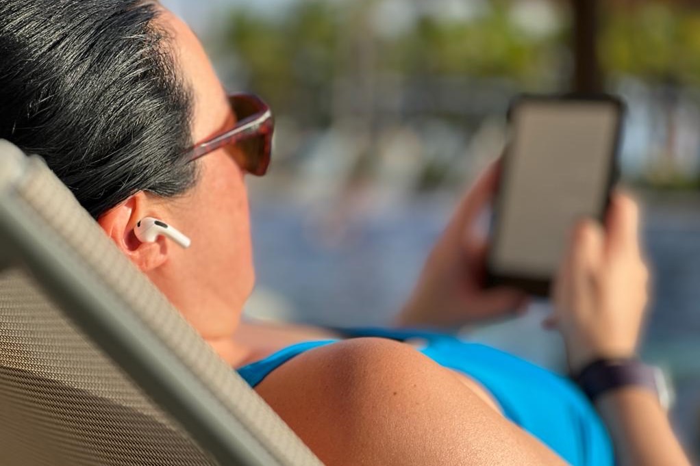 Frau am Pool mit Apple AirPods-Ohrhörern und einem E-Reader.