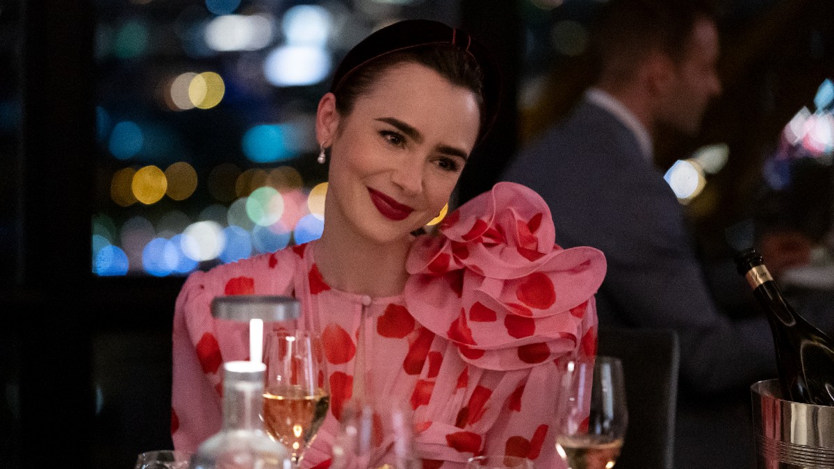 Lily collins smiles while sitting at a table from Emily in Paris.