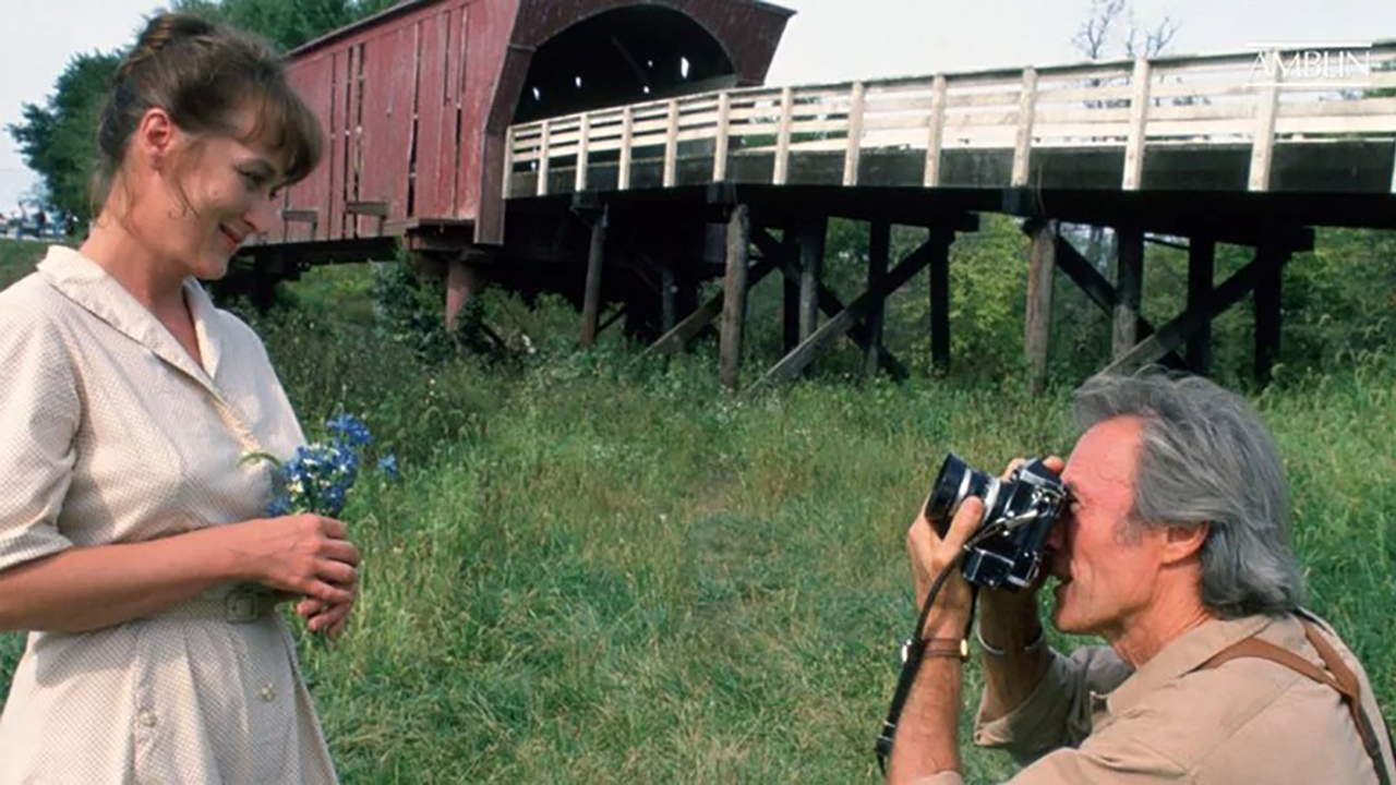 Meryl Streep posando para uma foto enquanto Clint Eastwood se ajoelha para tirar uma em The Bridges of Madison County.