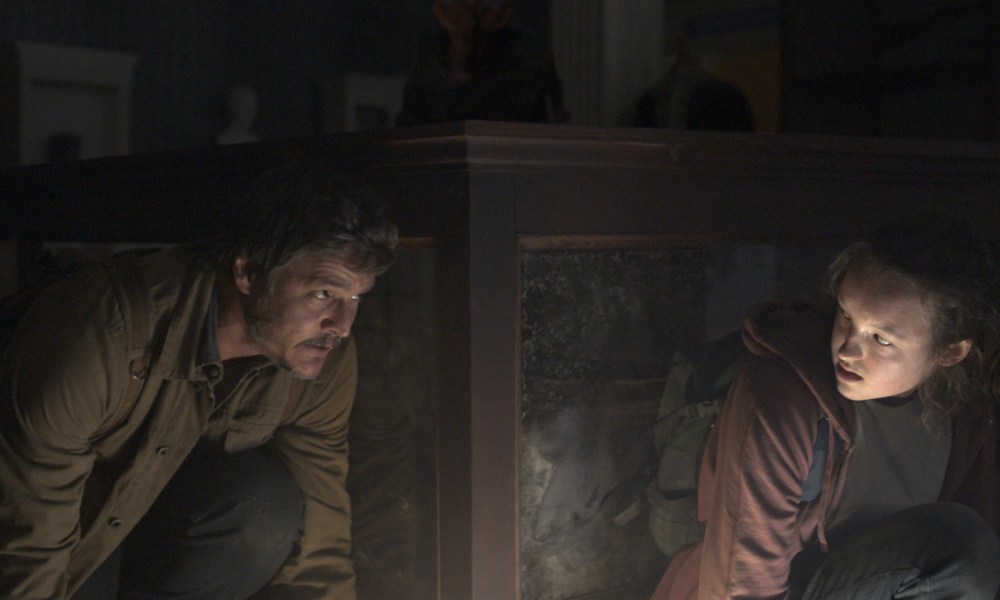 Pedro Pascal and Bella Ramsey crouch behind a glass case together in The Last of Us.