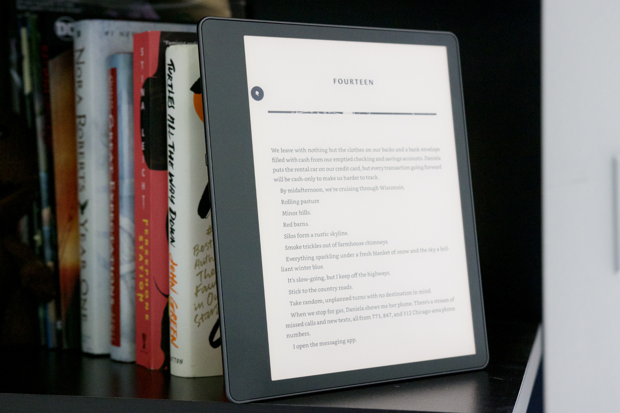 Amazon Kindle Scribe sentado en una estantería junto a una pila de libros.
