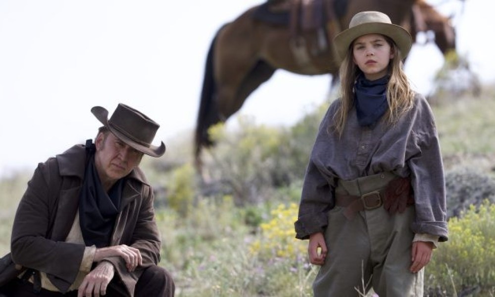 Nicolas Cage and Ryan Kiera Armstrong stare at the camera with a horse behind them in a scene from The Old Way.