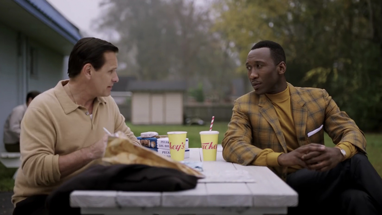 Deux hommes sont assis à une table de pique-nique dehors, mangeant et discutant dans une scène de Green Book.