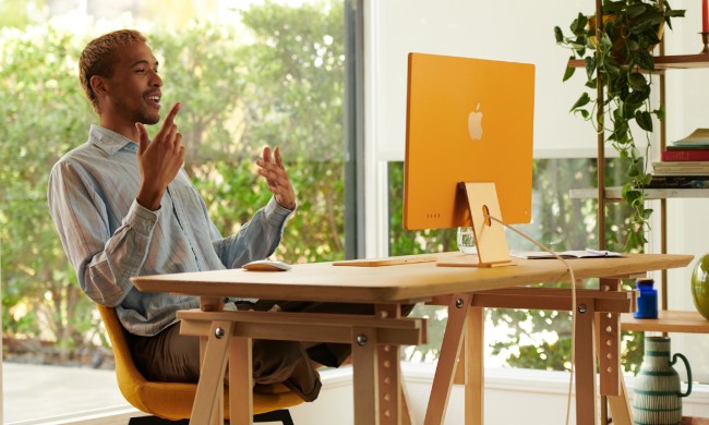 A man sitting at a desk in front of an M1 iMac. Behind him is a large glass window and a set of shelves holding books, plants and ornaments.