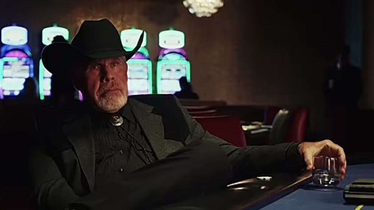 Ron Perlman avec un chapeau de cow-boy assis à une table de poker tenant un verre dans une scène de Poker Face on Peacock.