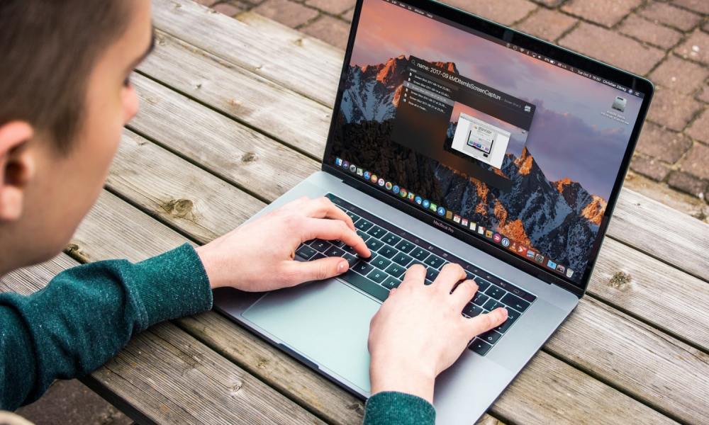 A person typing on a MacBook Pro while sat on a wooden bench.