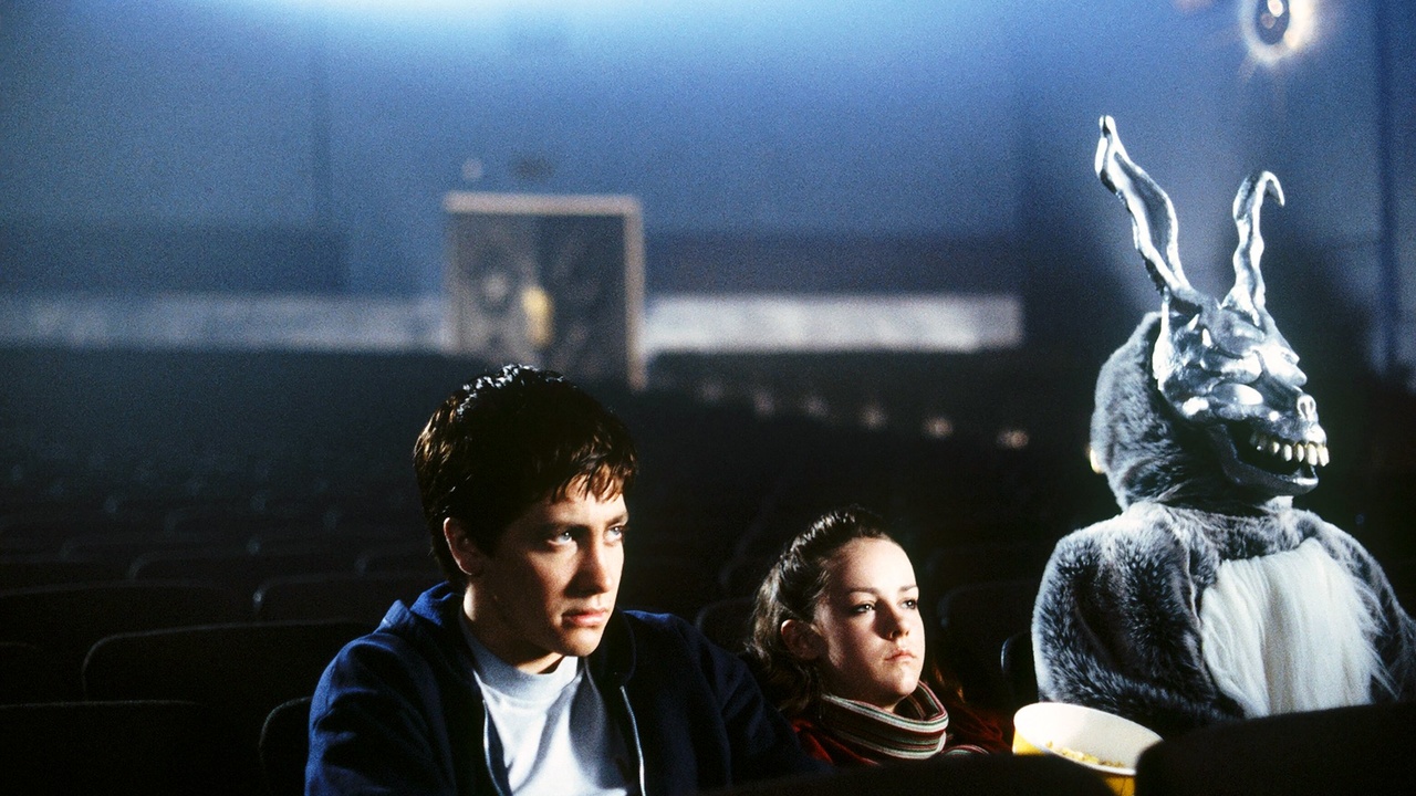 Jake Gyllenhaal e Jena Malone como Donnie e Gretchen no cinema sentados ao lado de um coelho gigante em Donnie Darko.