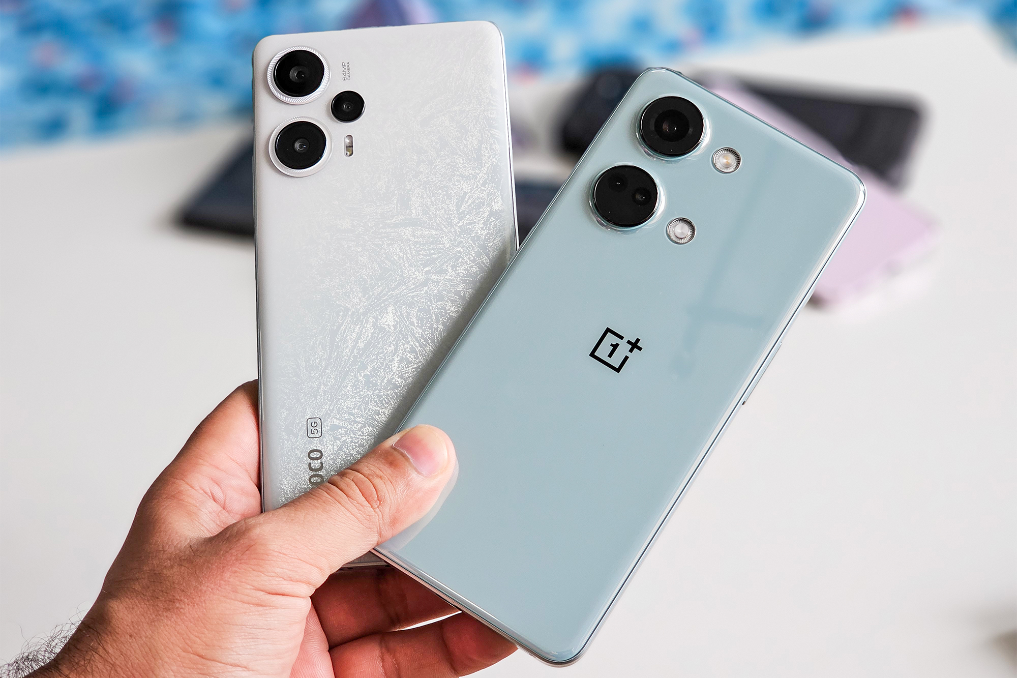 Xiaomi POCO F5 blanc et OnePlus Nord 3 vert bleuté tenus en main devant un tas de smartphones Android.