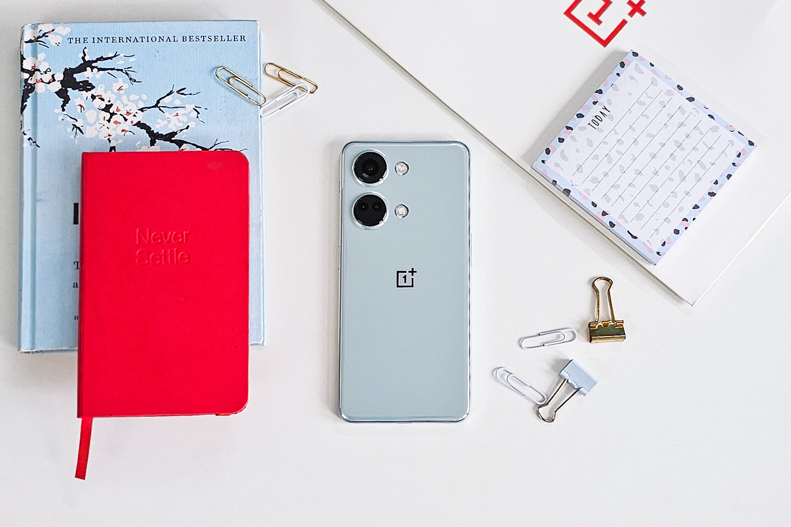 Color OnePlus Nord Misty Green sobre fondo blanco con un cuaderno Never Settle rojo, notas adhesivas, clips y un libro azul en el lateral.