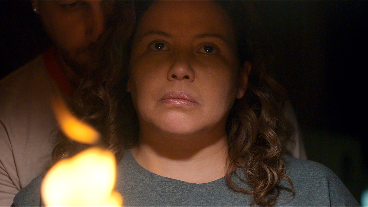 Justina Machado as Dolores Roach in The Horror of Dolores Roach, looking up in a dark room, a candle behind her.