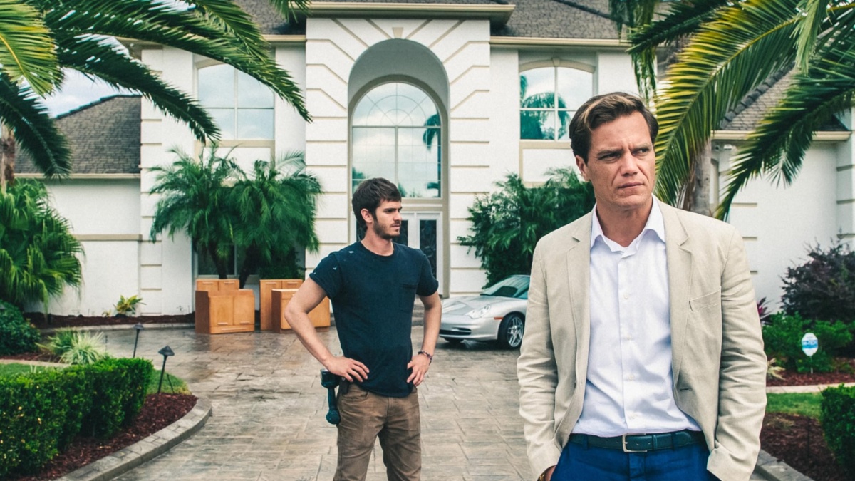 安德鲁·加菲尔德 (Andrew Garfield) 站在迈克尔·珊农 (Michael Shannon) 身后，站在 99 Homes 的一栋房子前。