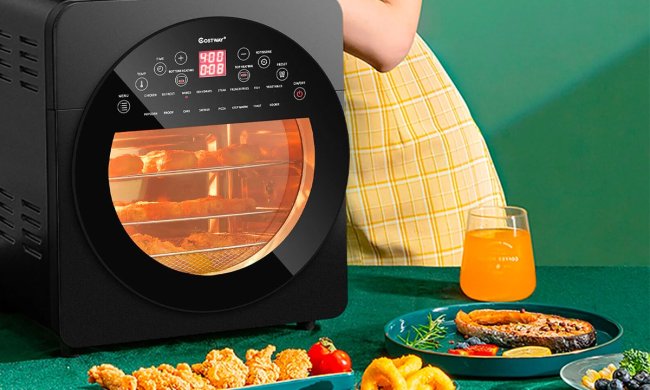 The Costway 16-in-1 15-quart air fryer oven on a table with plates of food spread out around it.