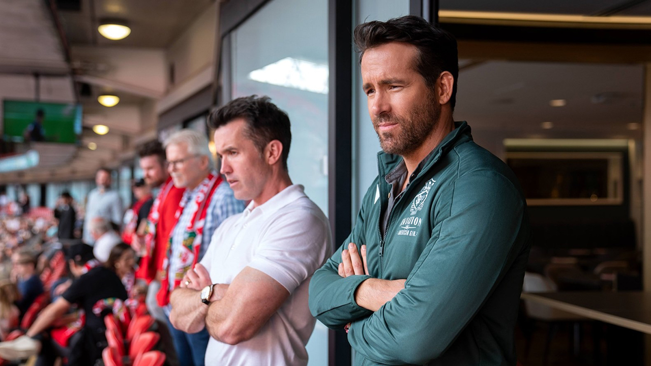 Rob McElheny e Ryan Reynolds assistem a um jogo de futebol em uma cena de Welcome to Wrexham