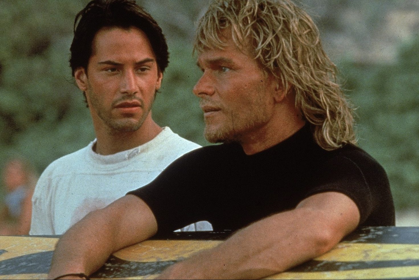 Un hombre sostiene una tabla de surf junto a otra persona en Point Break.