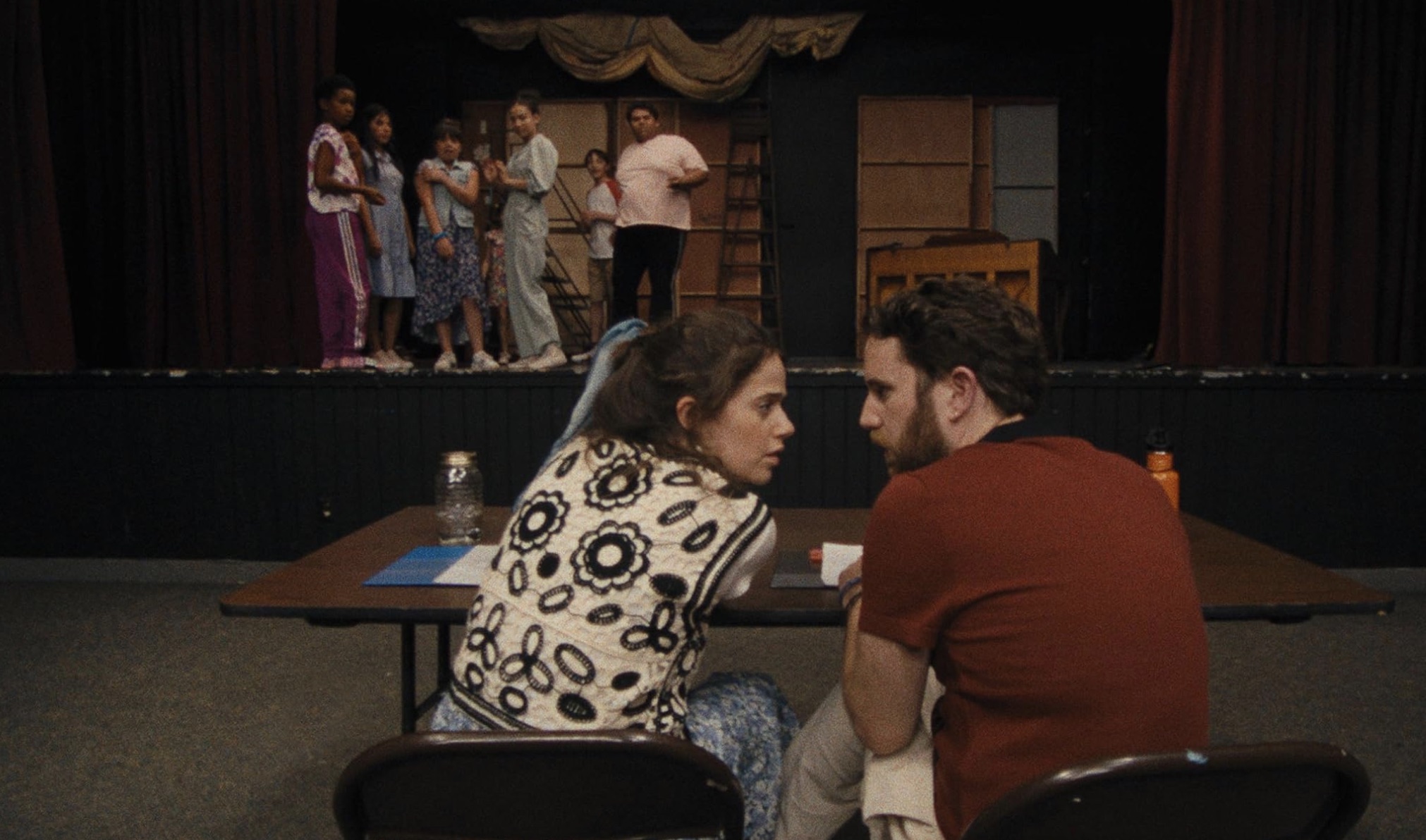 Dois adultos sentam-se à mesa enquanto as crianças sobem no palco do Theatre Camp.