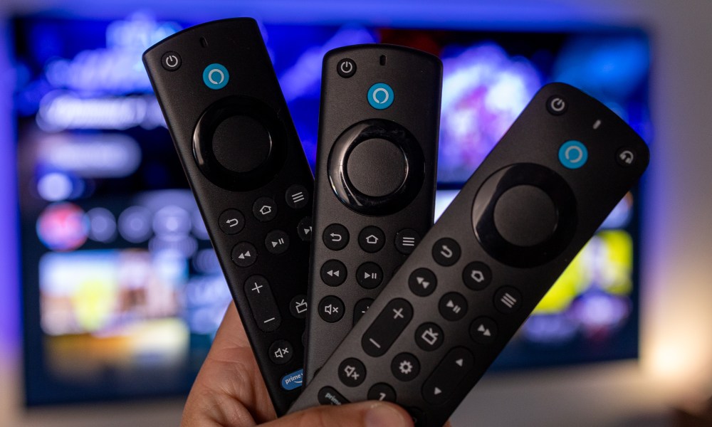 A trio of Amazon Fire TV remote controls held in a hand in front of a television.