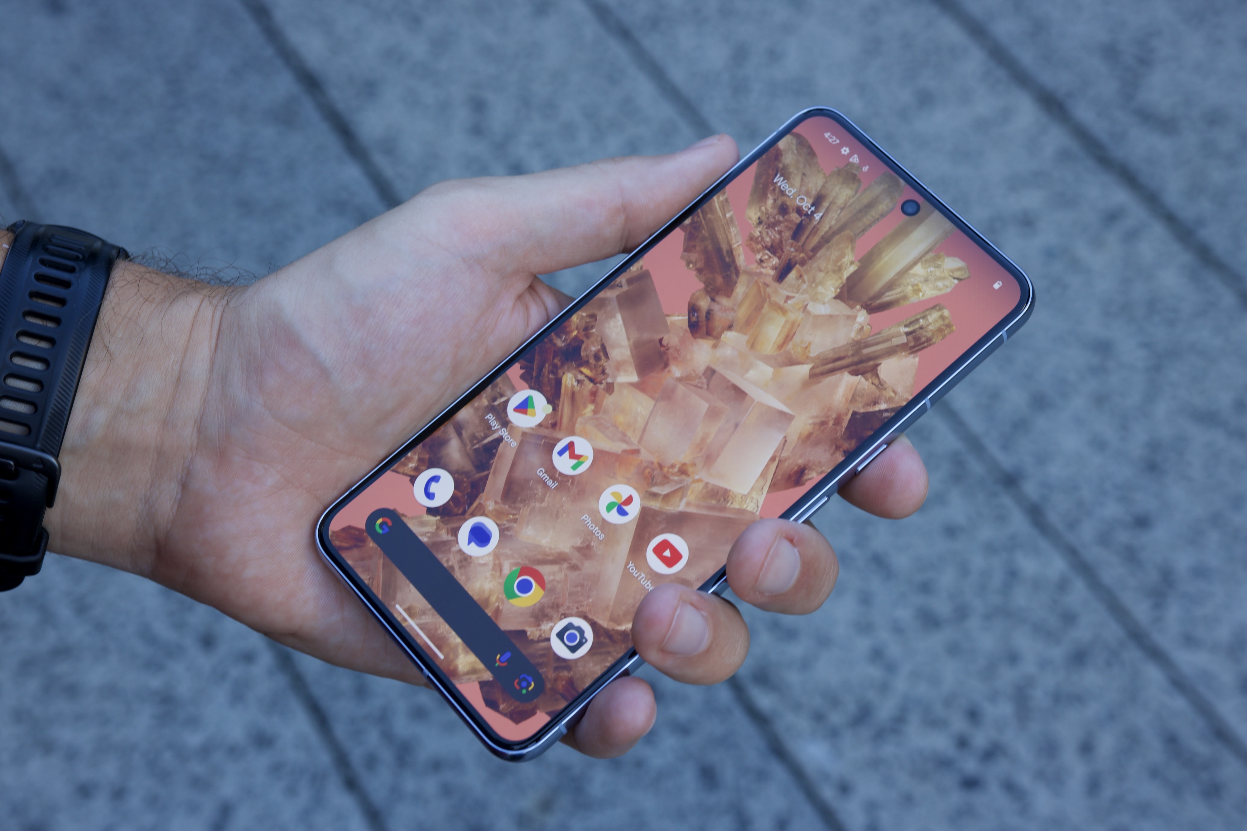 Holding a Google Pixel 8 Pro, showing its Home Screen.
