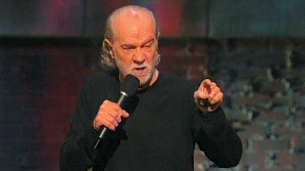 George Carlin sur scène avec un micro pointant un doigt et grimaçant dans une scène de George Carlin : You Are All Diseased.