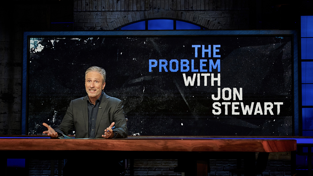 Jon Stewart sitting behind a large desk, hands turned up and a surprised expression on his face in The Problem With Jon Stewart.