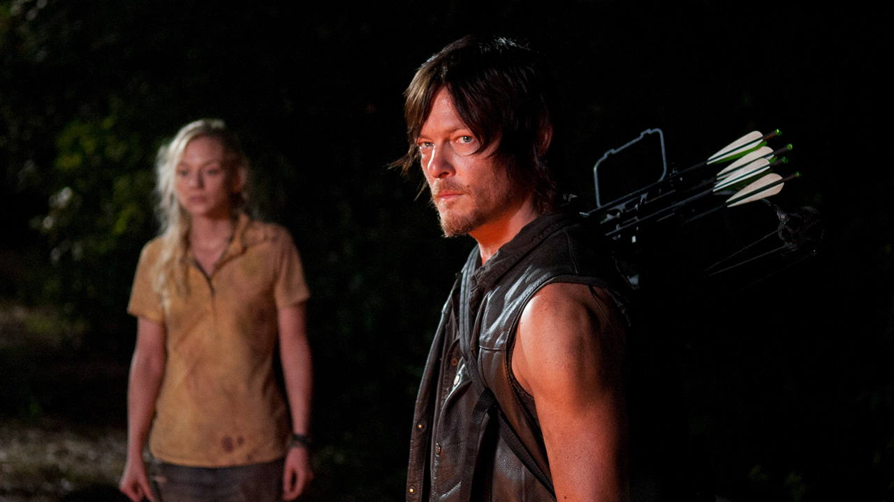 Daryl em pé, parecendo ameaçador com Beth ao fundo em uma cena de The Walking Dead.