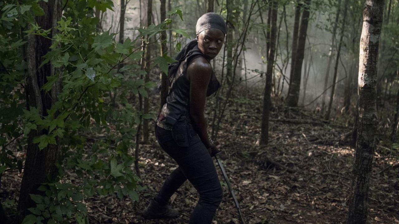 Michonne wearing a head scarf, standing in the middle of the forest with a weapon in a scene from The Walking Dead.