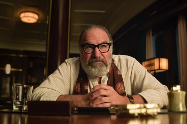 Mandy Patinkin sitting at a desk, hands clasped in front of him in a scene from Death and Other Details.