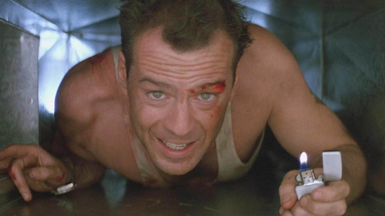 Bruce Willis como John McClane em Die Hard, rastejando por um respiradouro com o rosto ensanguentado e um isqueiro na mão.