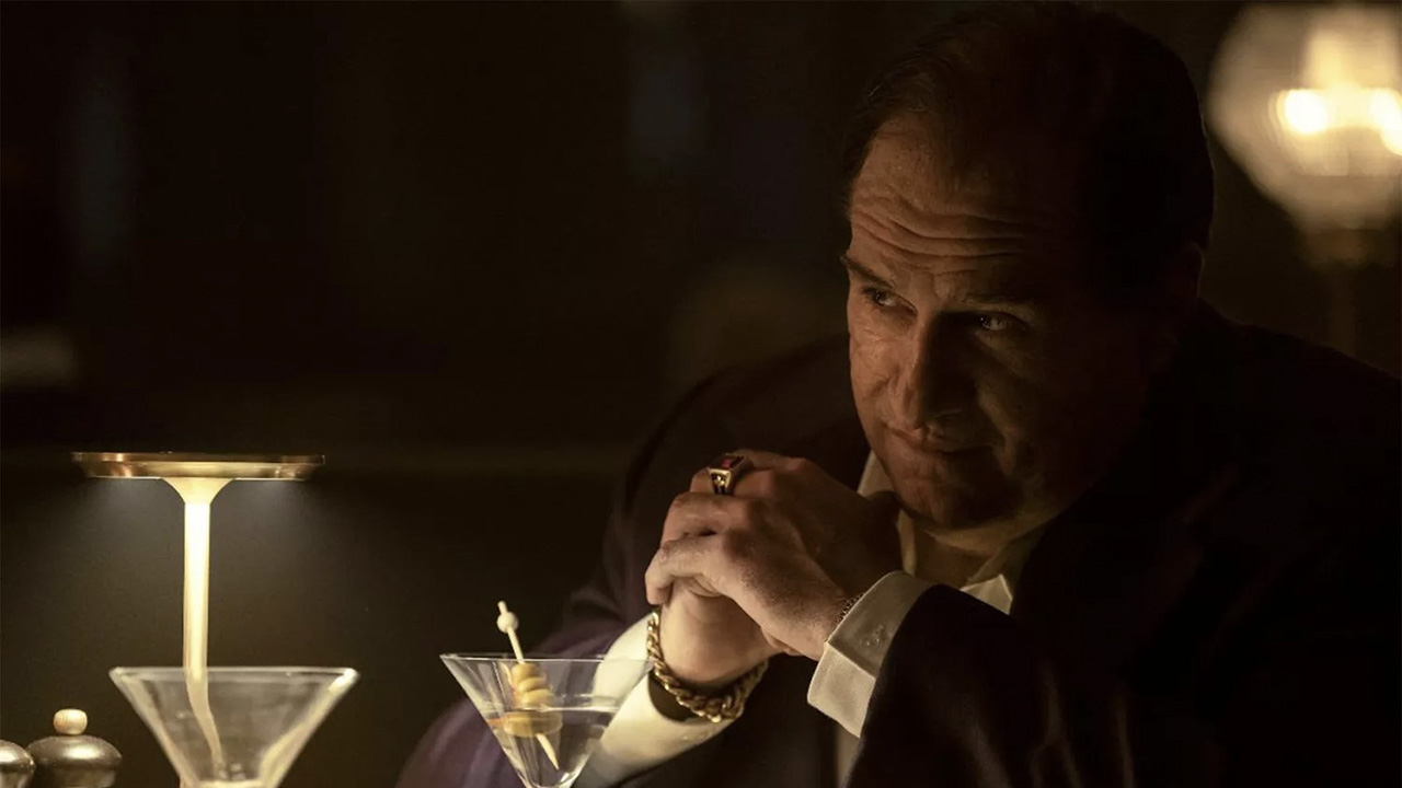 The Penguin sitting at a table in the dark, his hands clasped together in a scene from the series.