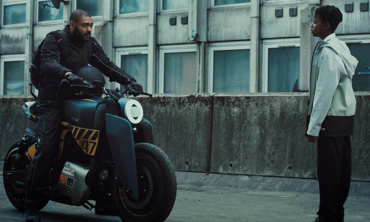 Izi sits on his motorcycle in front of Benji in The Kitchen.