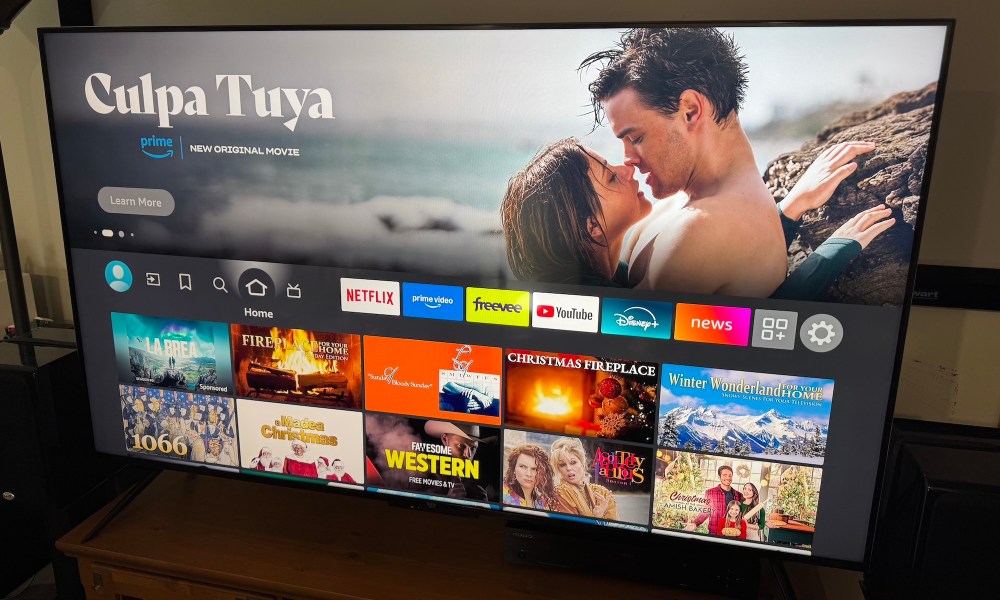 The Amazon Fire TV Omni Mini-LED TV displaying its home screen on a wooden home theater credenza.