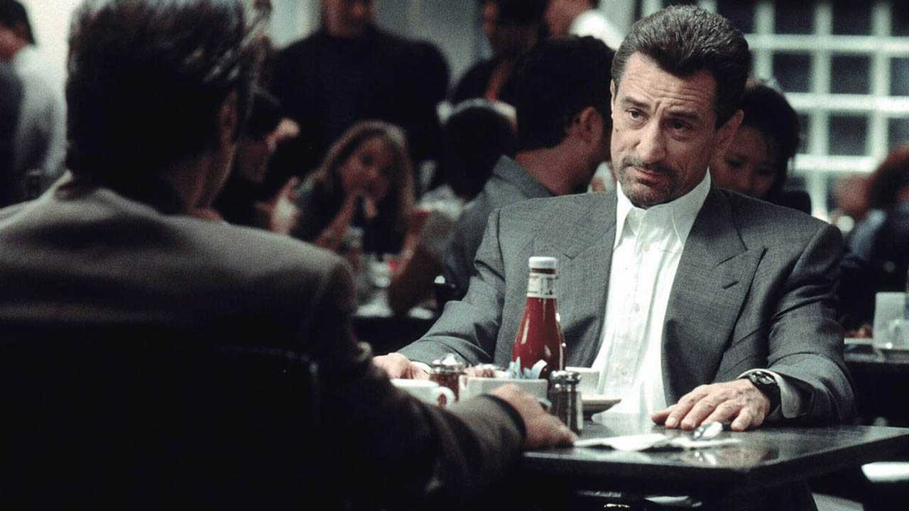 A young Robert De Niro sitting at a table in a restaurant with another man, looking unimpressed in a scene from Heat.