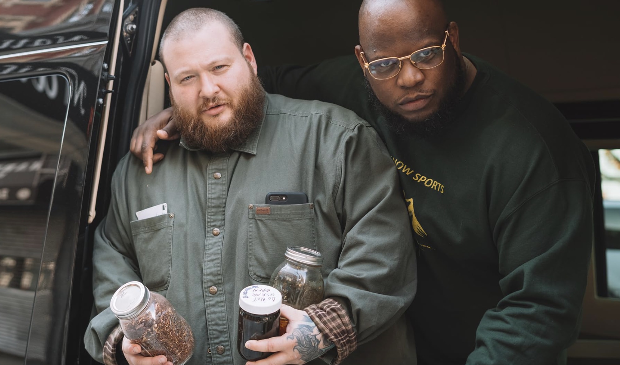 Action Bronson tient deux bocaux tandis qu'un autre homme pose sa main sur son épaule.