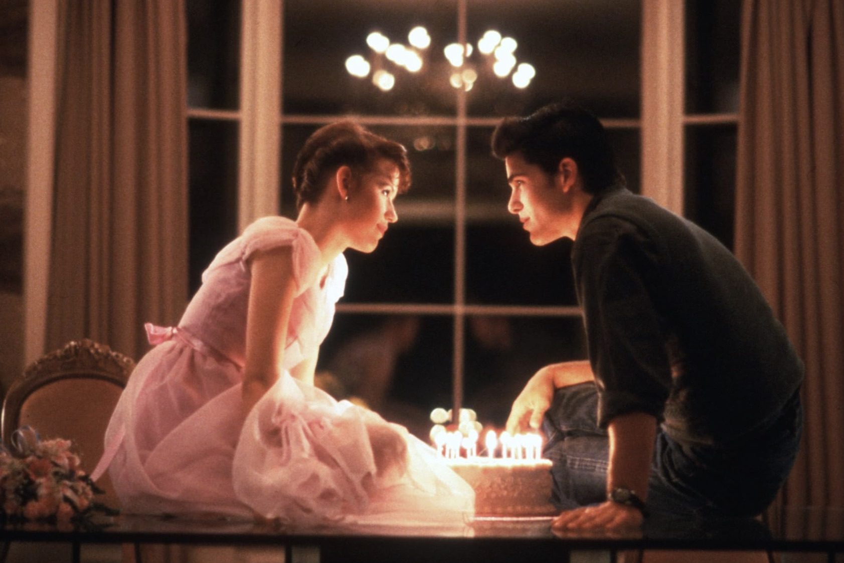 Molly Ringwald e Michael Schoeffling sentam-se em uma mesa e debruçam-se sobre um bolo de aniversário.