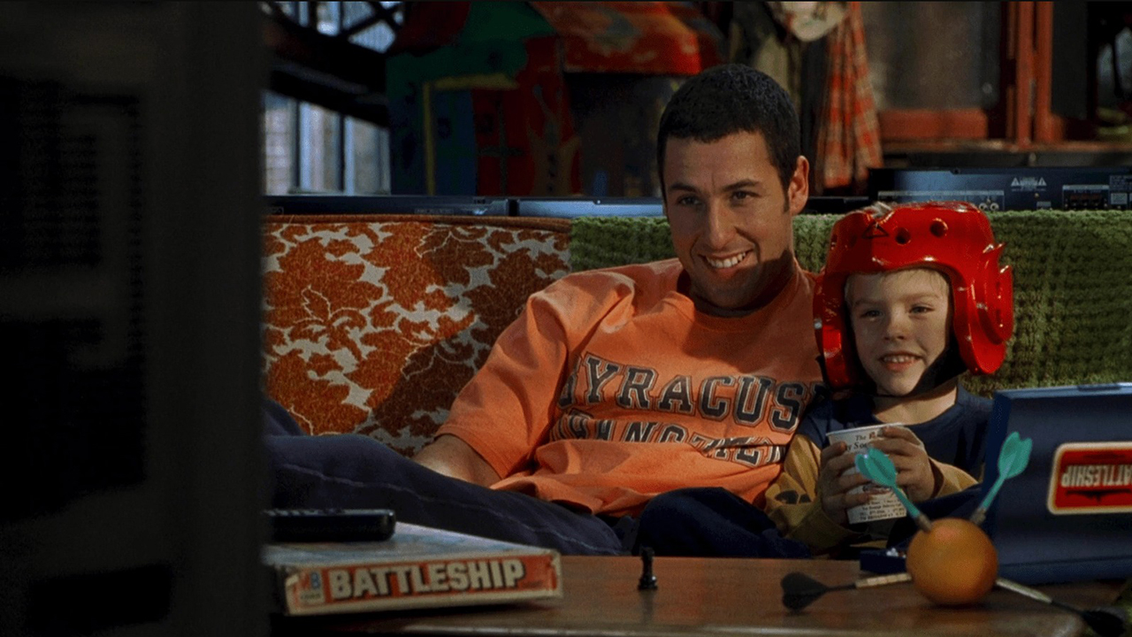 亚当·桑德勒 (Adam Sandler) 拥抱着一个戴着头盔的小男孩，两人在《老爹》(Big Daddy) 的场景中一起看电视。