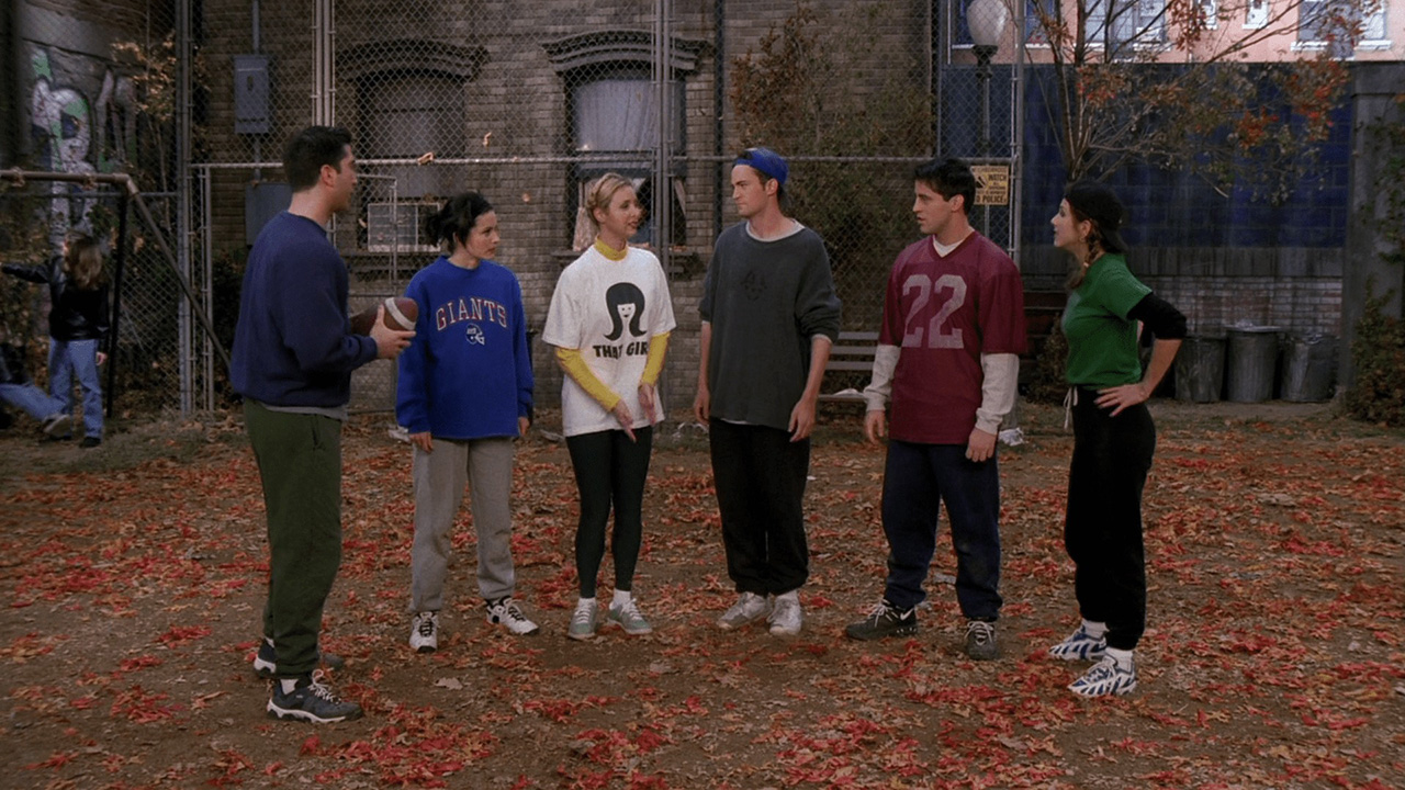 Ross, Monica, Phoebe, Chandler, Joey, and Rachel wearing football jerseys standing on the grass in a scene from Friends.