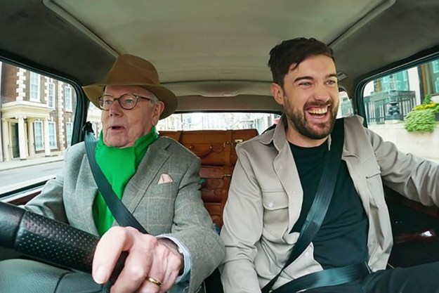 Jake Whitehall in a car with his father Michael driving, his father looking terrified and Jack smiling in a scene from Jack Whitehall: Travels with My Father on Netflix.